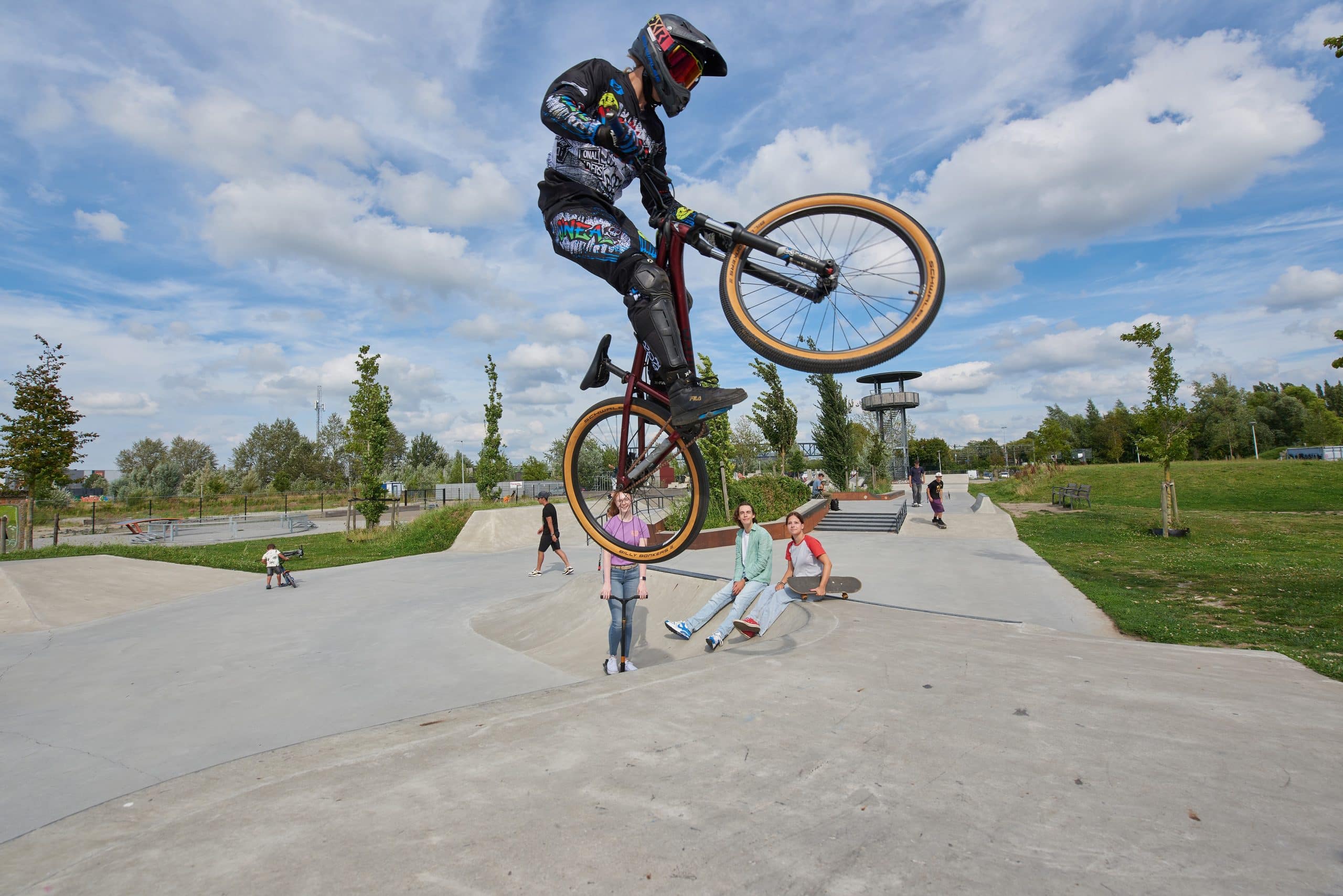 Een BMX-rijder springt hoog boven het Urban Sportspark in Roosendaal terwijl kinderen en tieners beneden kijken. De rijder draagt beschermende kleding en helm. Op de achtergrond liggen ramps, een groen veld en een paar mensen op skateboards en steps.