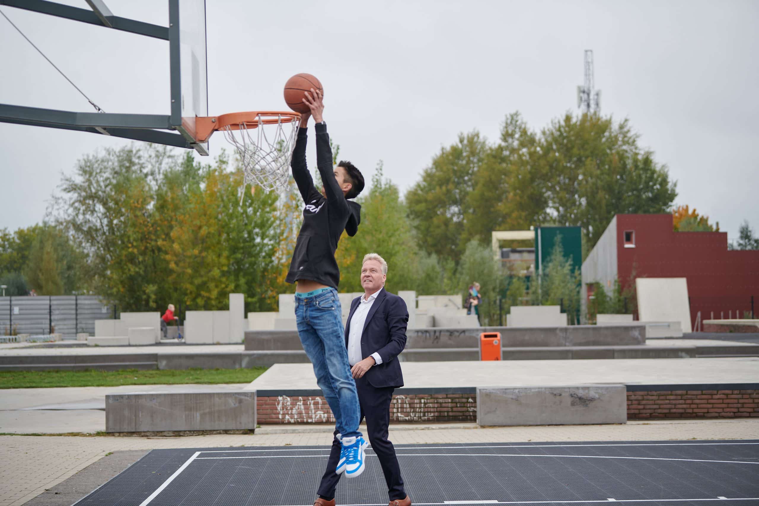 Een jongen dunkt een basketbal in het net, Burgemeester Buijs loopt langs